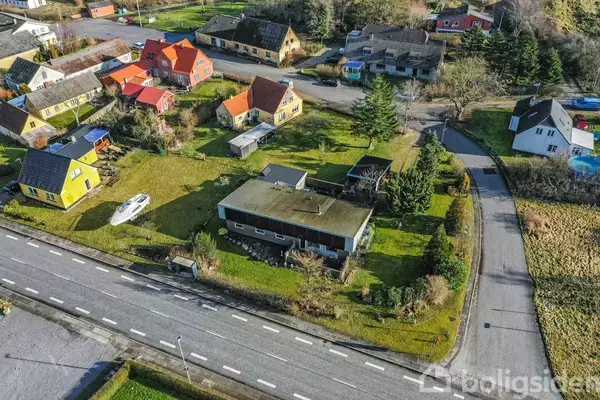 Et hus ses fra oven, omgivet af en have med træer og en båd på en trailer. Det er placeret nær en vej og andre huse i et kvarter.