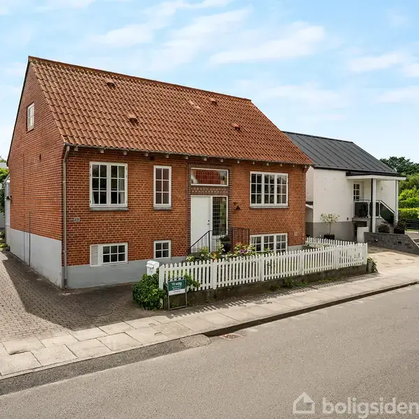Rødt murstenshus med skråt tag står stille på en fortovskant. Et hvidt stakit omkranser forhaven. Ved siden af er der en garage. Sollys skinner på en rolig gade.
