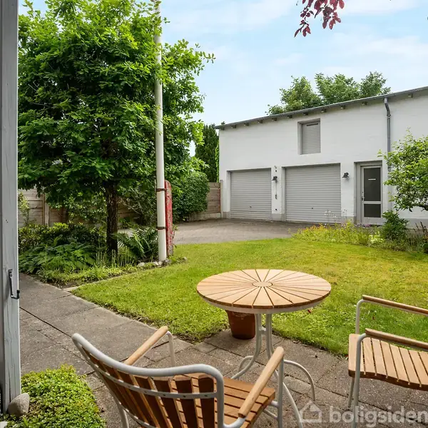 Terrasse med runde havemøbler foran græsplæne og træ. Til højre ses en lys garagebygning med to porte. Omgivet af buske og frodig vegetation.