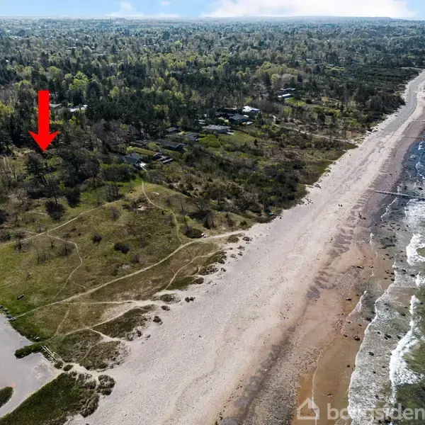 En rød pil peger mod et område ved en skov nær en strand. Stranden strækker sig langs en blågrøn havlinje, mens skoven omkranser en kystnær sti og et lille parkeringsområde.