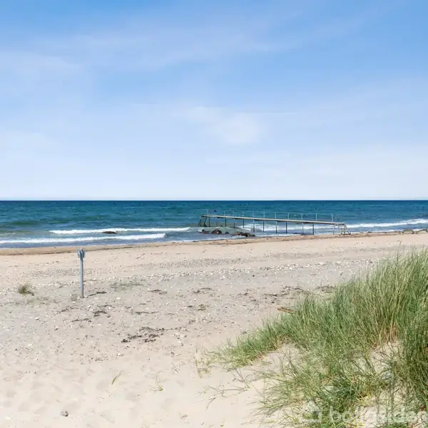 Strand med sand og græs i forgrunden. En badebro strækker sig ud over havet i baggrunden. Blå himmel og roligt vand kompletterer den fredfyldte scene.