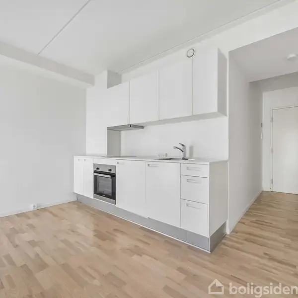 Et hvidt køkken med moderne skabe og indbyggede hvidevarer langs en væg. Trægulv dækker rummet, der har en åben indgang til en tilstødende gang.