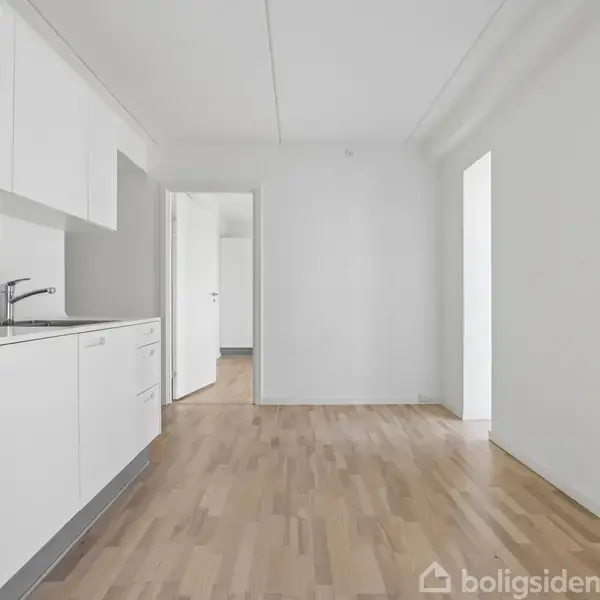 Et moderne, hvidt køkken med integrerede apparater og vask. Trægulv strækker sig ind i en åben stue. Lyse vægge og lofter skaber en rummelig atmosfære. Ingen tekst til stede.