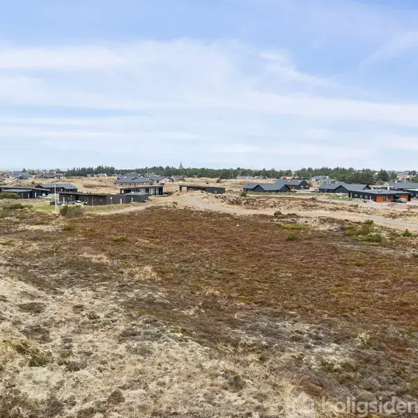 Huse ligger stille på en vidtstrakt, tør sandhedejord. Baggrund med flere beskedne boliger, omgivet af spredt vegetation under en klar himmel. Kontext: boliger i et naturlandskab.