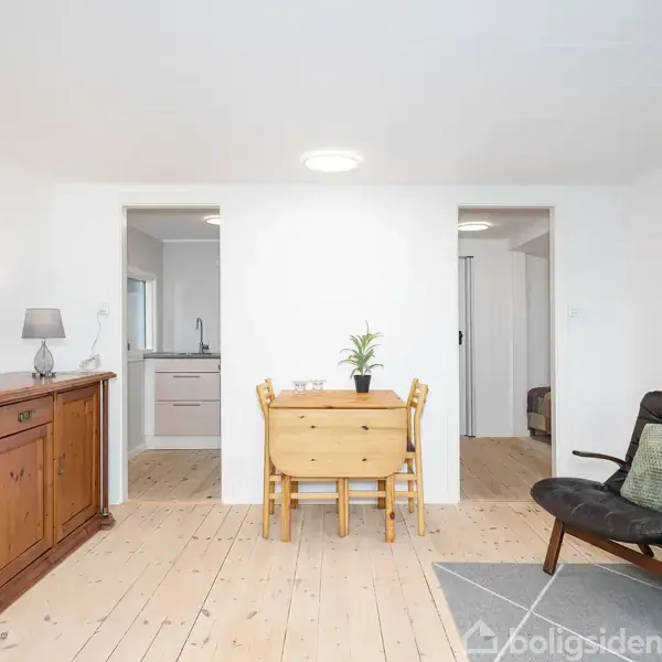 Et lyst rum med en trækommode til venstre og et lille spisebord med to stole i midten. Til højre er der en lænestol. To døråbninger i baggrunden fører til andre rum.