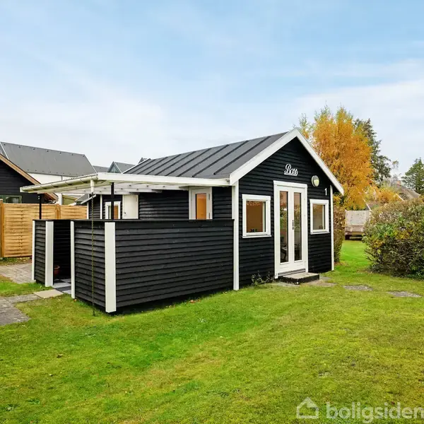 Et sort træsommerhus med hvide detaljer står i en velplejet have med grønne plæner og en busket baggrund. Overdækket terrasse på siden med en flisebelagt sti foran huset.