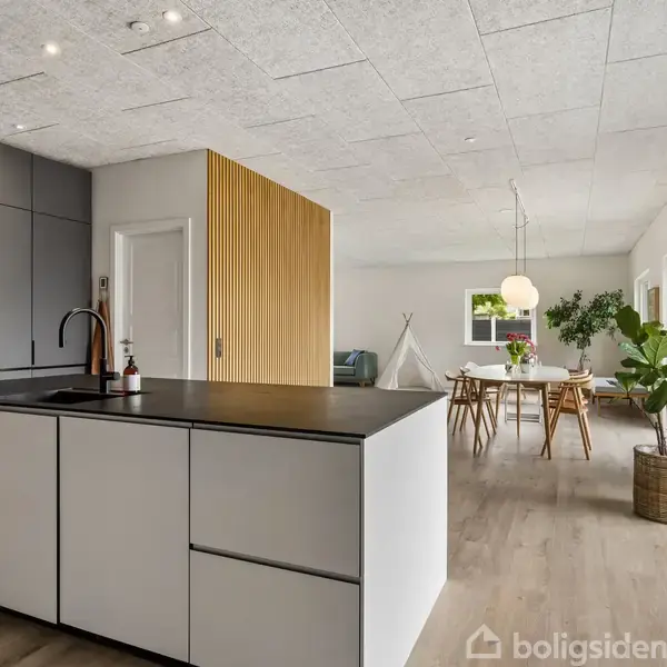 Køkken med hvid køkkenø og sort bordplade, stående i et åbent rum. Spisebord med stole og hængende lampe ses i baggrunden. Store vinduer giver lysindfald fra højre.