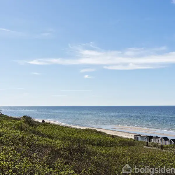 Kysten ligger roligt med små badehuse langs stranden. Havet strækker sig mod horisonten under en klar blå himmel, omgivet af grøn vegetation i forgrunden.