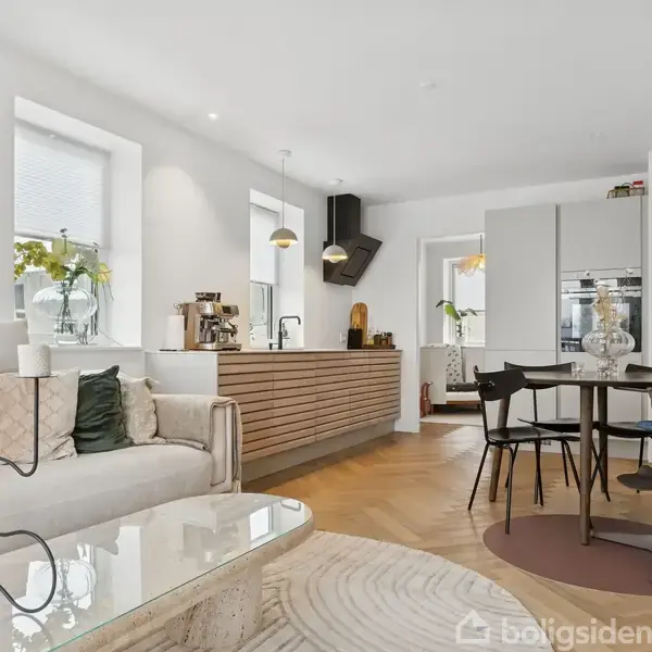 En moderne stue med en beige sofa, sofabord og åben køkken i baggrunden. Der er spisebord med stole, og lys falder ind fra vinduerne.