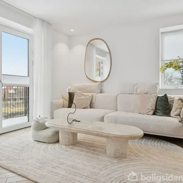 En lys stue med en beige sofa og et ovalt spejl på væggen. Et lavt, rektangulært sofabord står på et mønstret tæppe. Store vinduer slipper masser af lys ind.