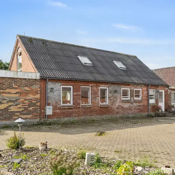 Et traditionelt murstenshus med skråt tag og tagvinduer står i en brolagt gårdsplads. Omkring huset er der planter og træer, der skaber en landlig atmosfære.