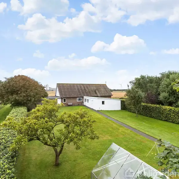 Et hus med stråtag står i en velplejet have med frugttræer og buske. En gennemsigtig struktur står i forgrunden, omgivet af marker og en blå himmel med skyer.