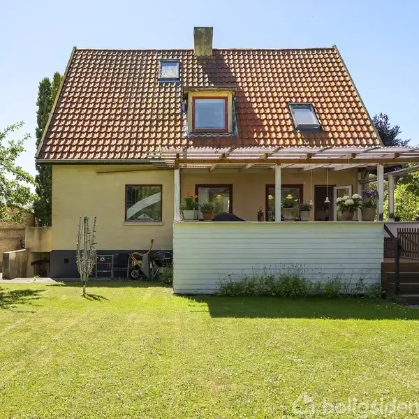 Et gulmalet hus med rødt tegltag står i en solrig have. En veranda med planter pryder facaden, og græsplænen omkranses af træer og buske.