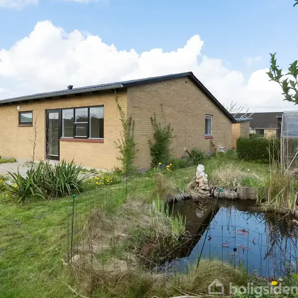 Et murstenshus stadig står stationært ved siden af en lille dam i en velplejet have indesluttet af buske, mens et drivhus ses i baggrunden under en skyfri himmel.