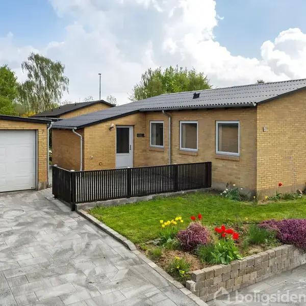 Et murstenshus med tilhørende garage ligger ved en flisebelagt indkørsel. Forhaven er anlagt med græs og blomsterbede, omgivet af et lavt, sort rækværk.