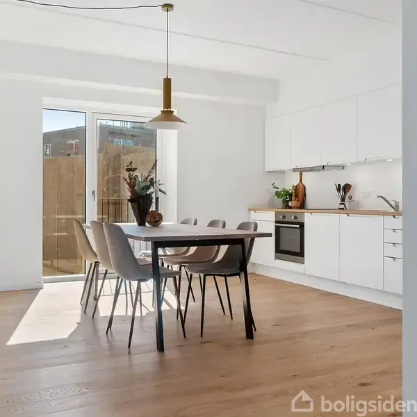 Et spisebord med seks stole står i et lyst køkken. Bordet har en vase med blomster. Bagved er der en moderne, hvid køkkenkrog med ovn, vask og skabe.