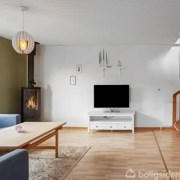 En stue med en blå sofa og sofabord til venstre, en sort brændeovn i hjørnet, en hvid tv-bænk med et fladskærms-tv foran en trappe til højre.