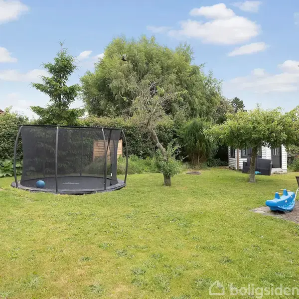 En trampolin står stationær på en velplejet græsplæne i en have. Til højre er et havebord med stole og en blå gyngehest. Omkring er der buske og træer.