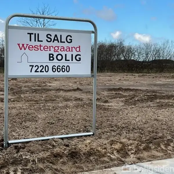 Et skilt står på en åben jordgrund. Skiltet siger: "TIL SALG Westergaard BOLIG 7220 6660". I baggrunden ses træer og en blå himmel med skyer.