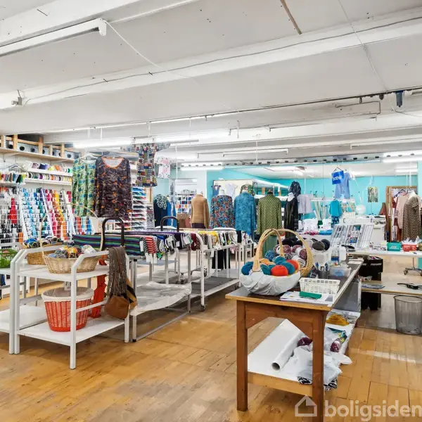Butik med skrædderartikler, inklusive tekstiler og syudstyr, organiseret på hylder og borde. Butikken har et farverigt udvalg af produkter, der skaber en kreativ og inspirerende atmosfære.