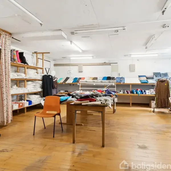 Butik viser tøj og tekstiler; borde med farverige stoffer midt i rummeligt lokale med trægulv. Mannequiner og hylder med ruller af stof i baggrunden, lyst op af loftslamper.