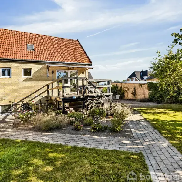 Hus med rødt tegltag og terrasse med trapper, beliggende i en have med plæne og brostenssti. Blomsterbede omkranser terrassen, og et træ står til højre.