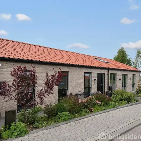 Et rækkehus med rødt tegltag står foran en flisebelagt fortov. Omkring huset er der grønt løv og små buske, mens himlen er blå med spredte skyer.