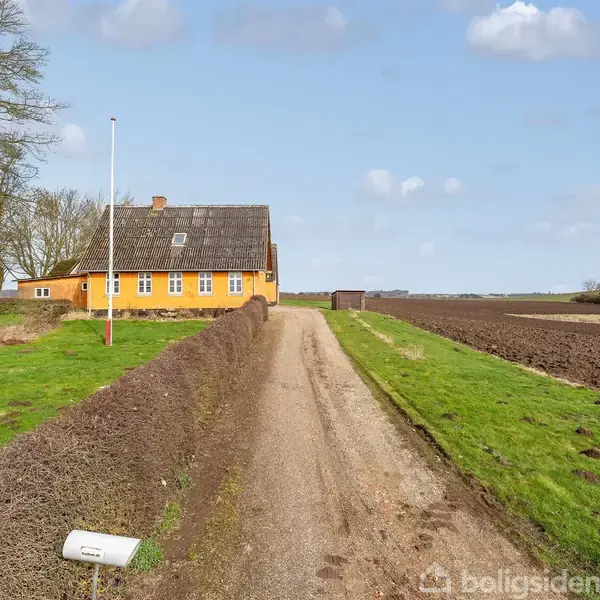 Et gult hus med stråtag står ved en grusvej omgivet af marker og træer under en blå himmel. En flagstang er placeret ved siden af huset.