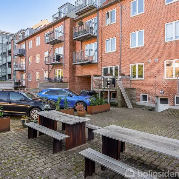 Bænke og borde står på en brostensbelagt gårdsplads. To biler er parkeret tæt ved en murstensbygning med altaner. Der er planter i kasser langs pladsen.