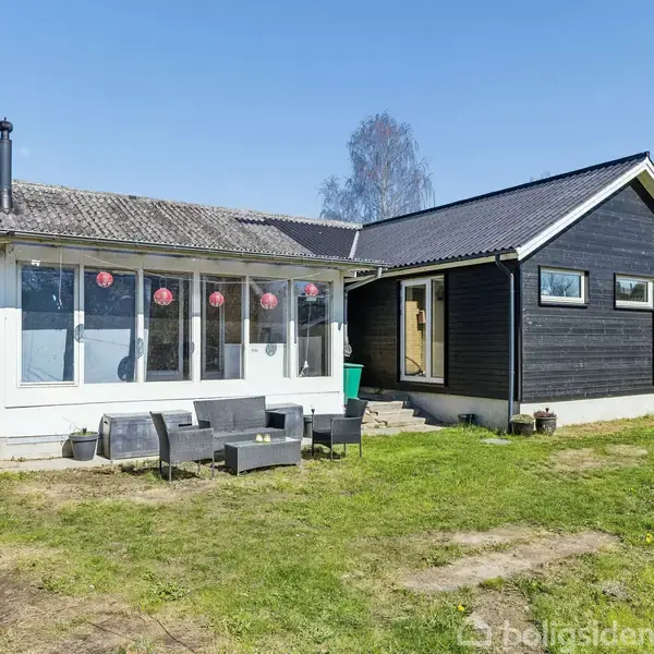 Et moderne sort-og-hvidt hus med store vinduer står stille i en frodig have. Havemøbler er placeret foran huset, og himlen er klar blå.