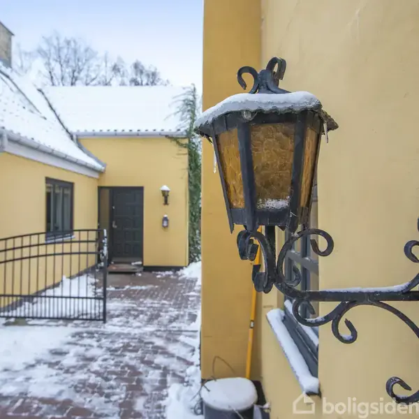 En væglampe dækket af sne hænger på en gul mur. Der er en snedækket indkørsel foran en port og hus med skråtag i baggrunden.