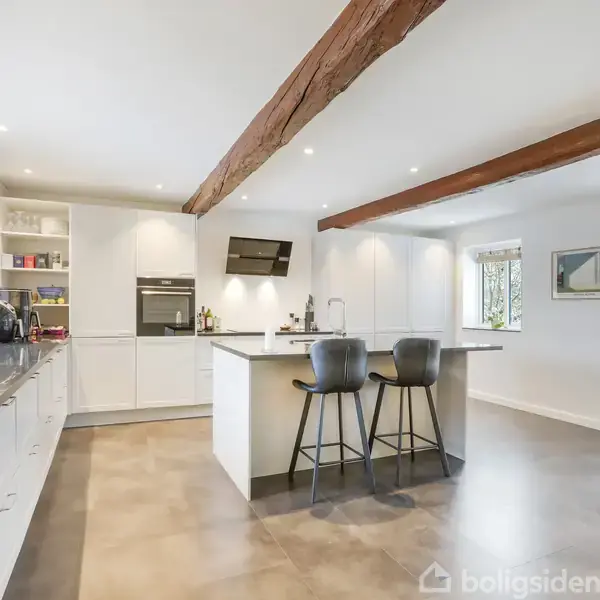 Køkken med hvidt interiør og en central ø med to sorte barstole. Ovenpå ses synlige træbjælker. Store vinduer giver udsigt til grønne træer. Komfur og væghylder ses i baggrunden.