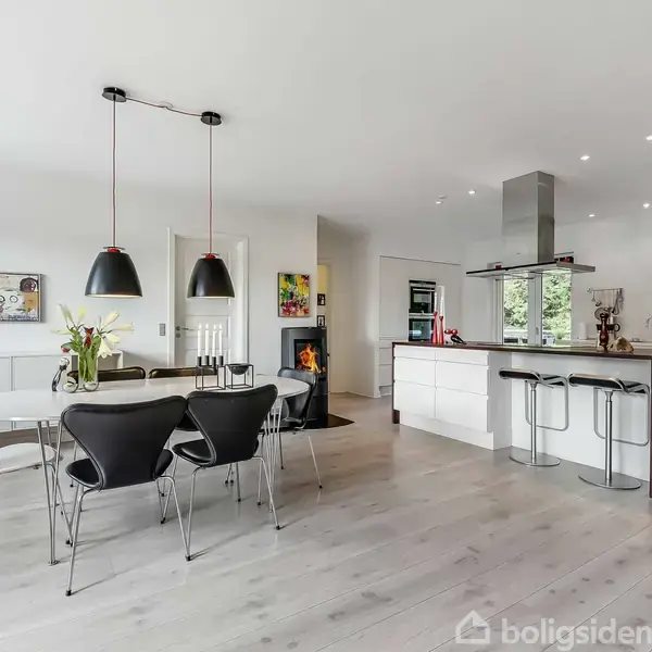 Et moderne køkken-alrum med hvidt interiør, en stor ø med barstole og et spisebord med sorte stole. Kunst på væggene og store, sorte loftslamper.