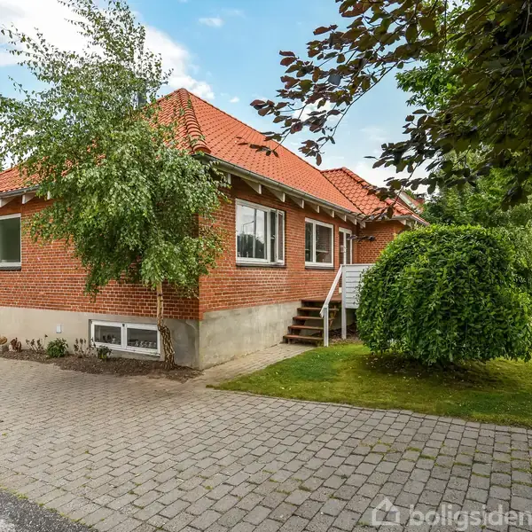 Et rødt murstenshus med et rødt tegltag står på en brolagt indkørsel, omgivet af buske og træer, i et frodigt grønt område.