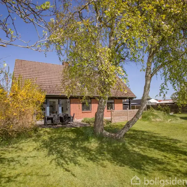 Et rødt murstenshus med brun tag ligger i en solrig have. Foran huset står træer med grønne blade. En terrasse med havemøbler ses ved husets indgang.
