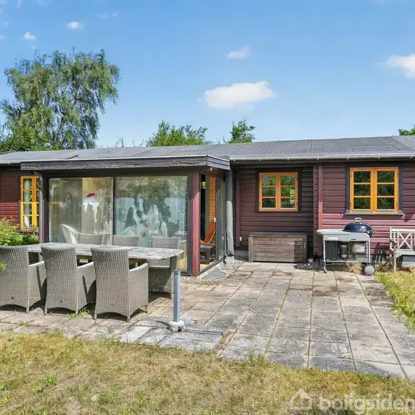 Hus med brun facade og gule vinduer står i baghaven. Terrassen har spisebord med fem flettede stole. Græs, buske og træer omgiver området, og himlen er klar med få skyer.