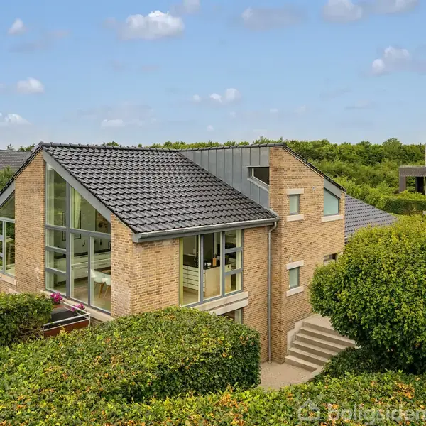 Et moderne hus i mursten står omgivet af velplejede hække. Huset har store glasfacader og en mørk tagkonstruktion. Andre huse er synlige i baggrunden under en blå himmel.