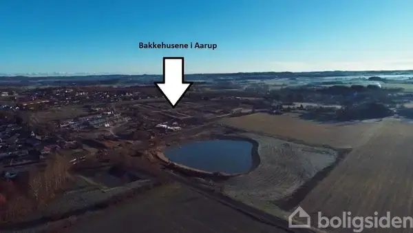 Pil peger mod byudviklingsprojekt "Bakkehusene i Aarup" i et åbent landskab med marker og et større vandhul under en klar, blå himmel.