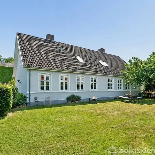 Hus med gråt tag og hvide vinduer. Det står stille i en velplejet have med grøn græsplæne og træer i baggrunden. Det er solskin og himlen er blå.