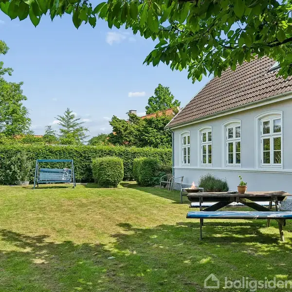 Et hvidt hus står i en velplejet have. En blå havesofa og et træbord med bænke er omgivet af grønne buske og træer under en blå himmel.