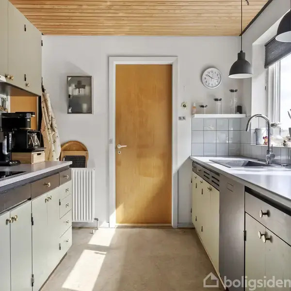 En køkkenindgang med en trædør i midten, omgivet af gulgrønne skabe og en mørk bordplade. Til højre ses en vask ved et vindue, med en vægklokke ovenover.