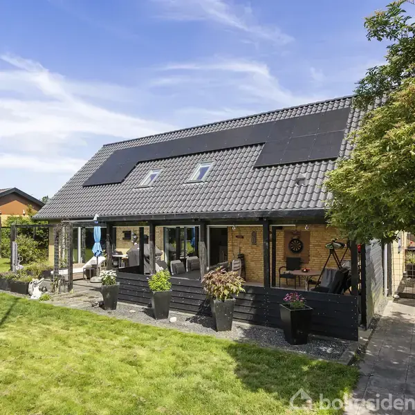 Hus med sorte solpaneler på taget står midt i en grøn have. Terrassen er delvist overdækket, og omgivende træer kaster skygger. Skyfri himmel i baggrunden.