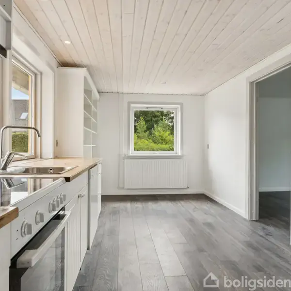 Køkken med hvide skabe og træbordplader. Komfur står langs væggen under et vindue, der lukker naturligt lys ind. Rummet har gråt gulv og træloft.