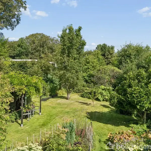 En frodig have med frodige træer og buske omkranser en græsplæne. En pergola med klatreplanter står til venstre, mens højbede ses i forgrunden.