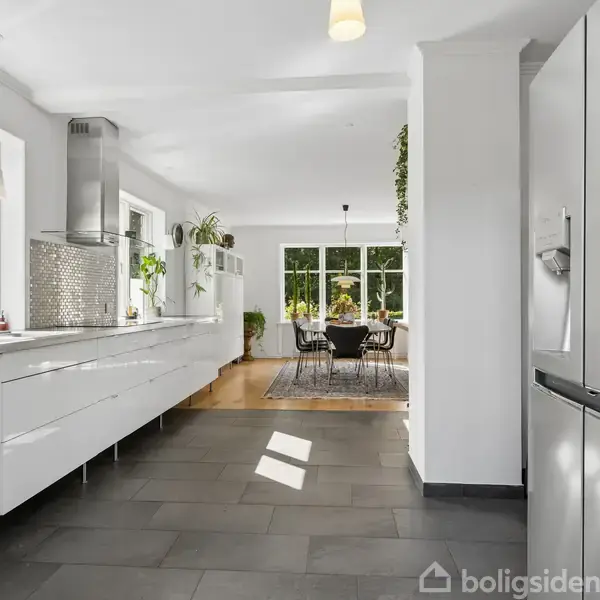 Køkken med hvide skabe og grå fliser, køleskab til højre. Lyst og rummeligt med planter på bordpladen. I baggrunden ses en spisestue med stort vindue og spisebord.