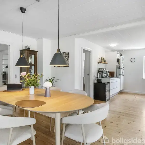 Et rundt spisebord med seks stole er placeret i et lyst køkkenalrum. Bordet er dekoreret med blomster, mens lamper hænger over. Køkkenet har hvide skabe og et stort vindue.