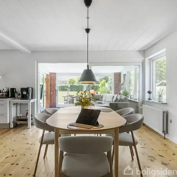 Et rundt spisebord med seks stole står i en lys spisestue. Bordet har en bog og vase. Rummet er åbent til en stue med store vinduer. En verdenskort hænger på væggen.