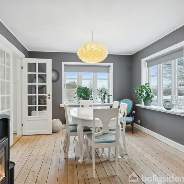 En spisestue med et rundt, hvidt bord omgivet af stole, opstillet på et trægulv. Et brændende brændeovn står i rummet, der har grå vægge og vinduer med planter.