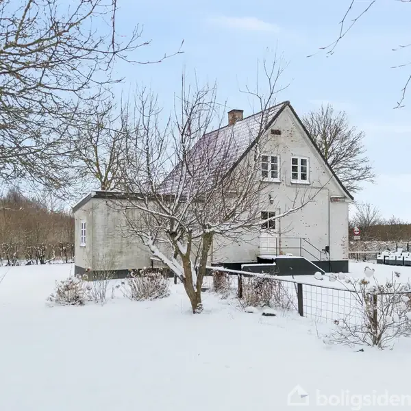 Et hvidt hus med mørkt tag står i en snedækket have. I forgrunden ses nøgne træer og en metaltrappe ved siden af huset. Landskabet er omkranset af træer.