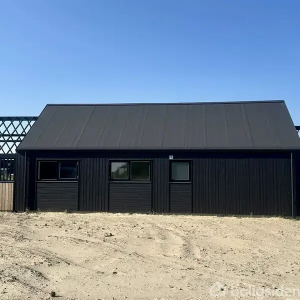 Sort hus med skråtag står på bar, sandet jord. Til venstre og højre for huset er der åbne træstrukturer med gitterdesign, og himlen er klar og blå.